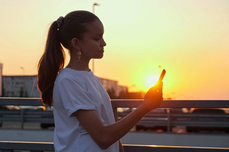 Schoolgirl looks into smartphone at sunset. Addicted young teenage girl with ponytail holds gadget in hand standing on bridge at back setting sun in summer