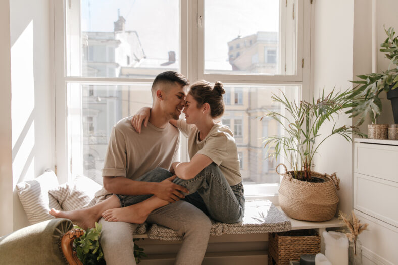 Cute young couple sitting on windowsill against city view background. Pretty girl with brunette collected hair in jeans and t-shirt relaxing near boyfriend in light clothes indoor