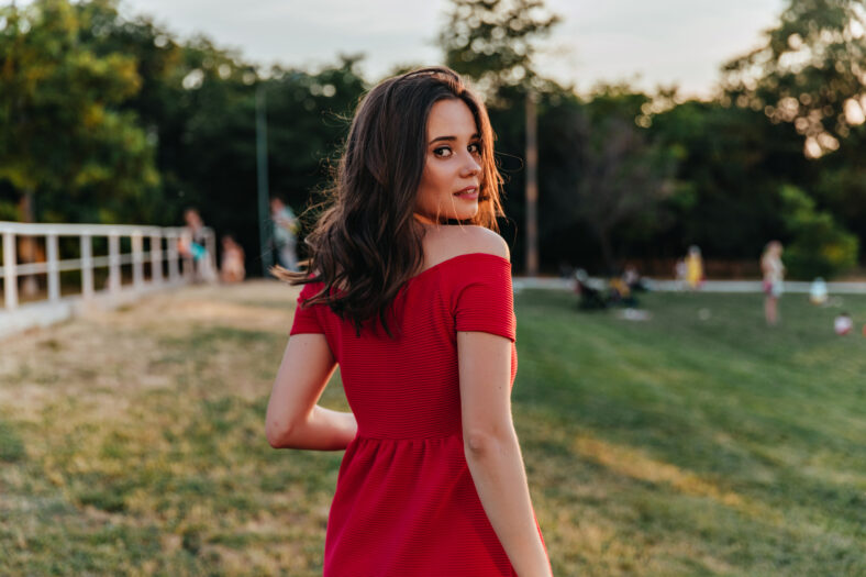 Dreamy brown-haired girl looking over shoulder while walking in park. Portrait of amazing caucasian lady in red dress standing on nature background.
