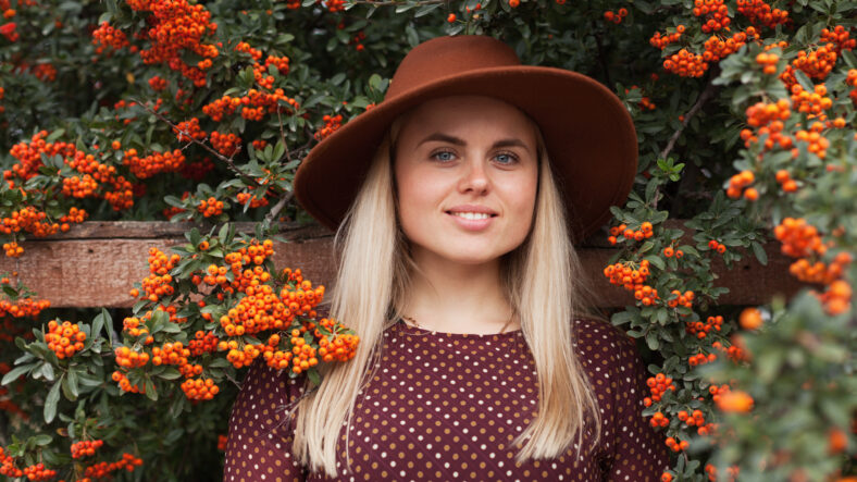 Beautiful woman wearing terracotta brown colous hat and rustic dress near rowan tree with ripe orange berries. Slow living and natural beauty concept.