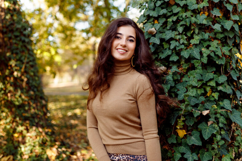 Beautiful autumn. Cheerful girl with long curly hair and makeup, wear in fall clothes, poses in park, near trees in day time.