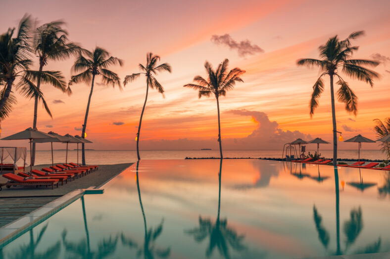 Fantastic poolside, sunset sky, palm trees reflection. Luxury tropical beach landscape, infinity swimming pool, deck chairs and loungers under umbrellas amazing scenic. Vacation resort hotel landscape