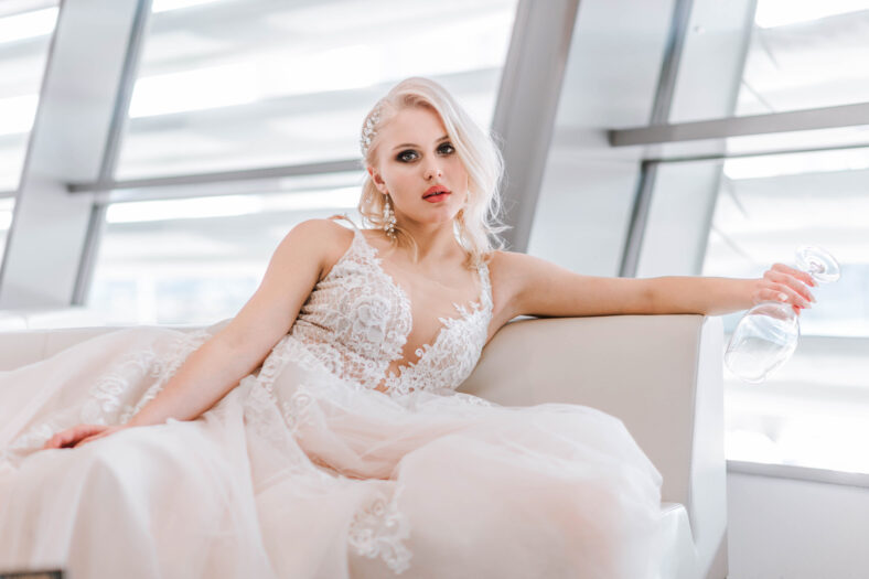 An attractive pensive blonde woman with a creative hairstyle for short hair is sitting on a white sofa in the lobby of the hotel. Art photography. White long wedding dress of the bride.