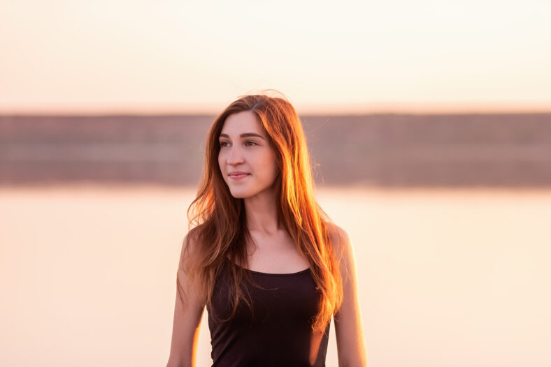 Retrato de primer plano de mujer joven con un vestido negro aireado contra el cielo al atardecer. El cabello de las chicas está iluminado por los rayos del sol. Una mujer camina por la orilla del mar al atardecer. Fin de semana de viaje de estilo de vida
