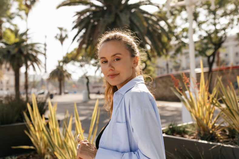 Close up outdoor photo of romantic pretty girl in blue shirt is posing at camera and enjoying summer walking. Smiling girl relaxes outdoors drinking coffee.