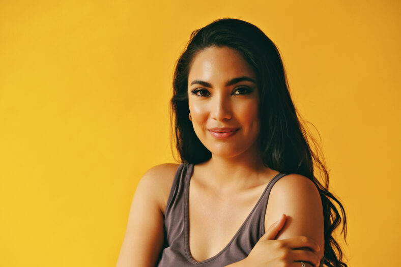 headshot beauty shot of sensual hispanic Latina woman smiling young adult with black long hair and tank top in front of yellow background looking at camera studio shot