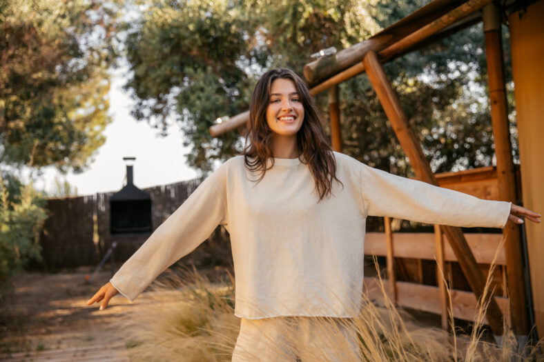 Positive young caucasian girl waving her arms walking in countryside during day. Brunette smiles broadly, wears spring clothes. Nature, summer holidays concept