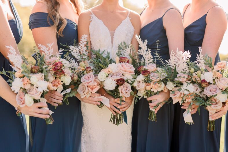 Bride and bridesmaids holding bouquets. Bridesmaids are wearing navy blue dresses. Bouquet has roses and wildflowers.