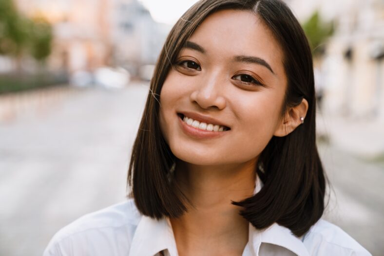 Brunette asian woman smiling and looking at camera while walking on city street