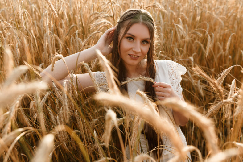 Portrait of young attractive female in dress standing in yellow wheat field and looking at camera