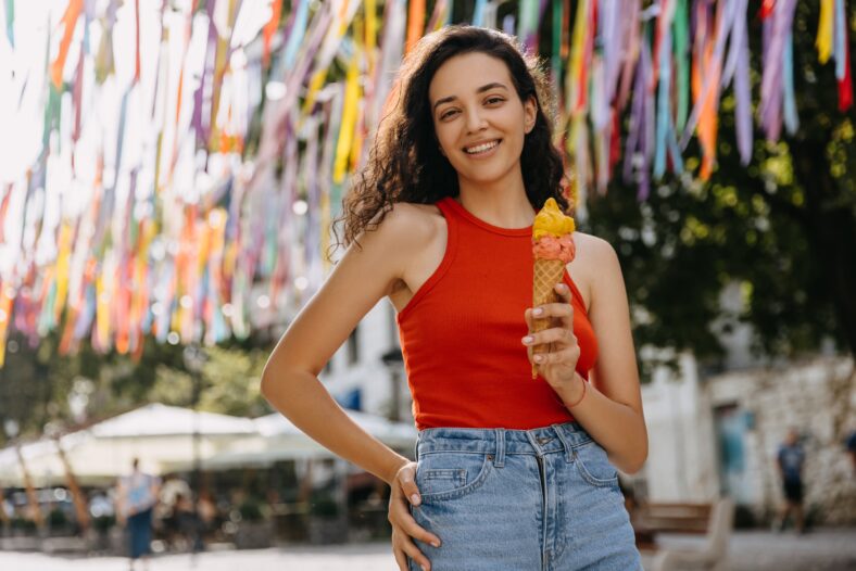 Happy young brunette woman on city street on a hot summer day, holding a big double layered ice cream.
