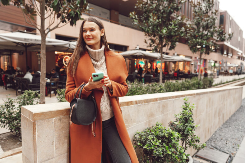 Young woman in brown coat using the phone on the city streets close up