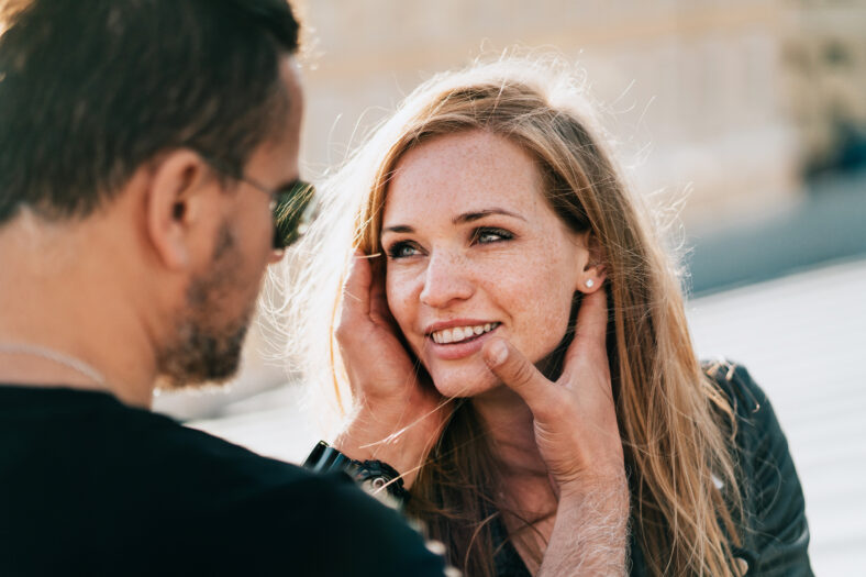 Close up of your adult American couple in black clothes looking at each other. Redhead cheerful caucasian girl looks tenderly at boyfriend. Romantic man touching wife’s face, date, relationship.