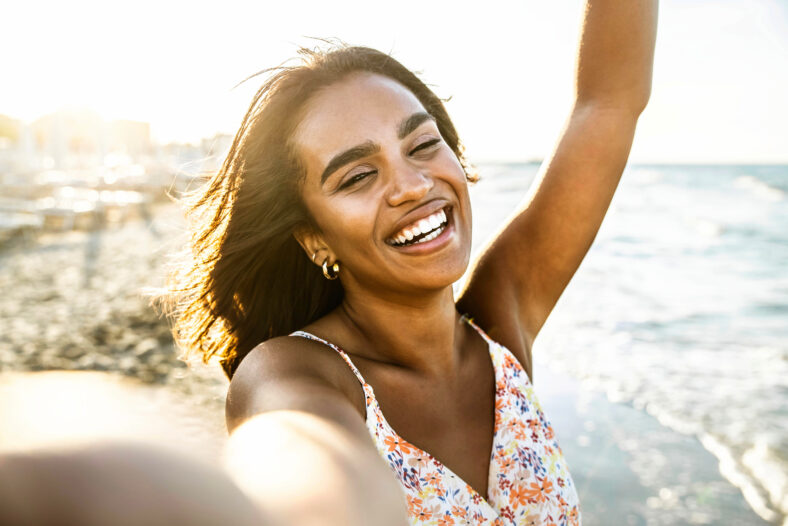 Hermosa mujer joven negra tomando una foto de selfie caminando en la playa - femenina encantadora sonriendo a la cámara afuera - vacaciones de verano, tecnología y concepto de estilo de vida saludable