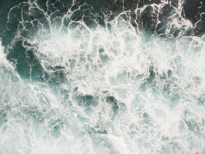 white sea foam on the ocean surface is agitated salt water from crashing waves and a strong surf, mixing currents and undertow swirl in the deep blue ocean during typhoon storms interrupt shipping