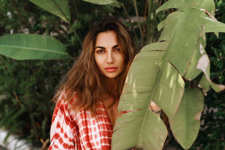Young woman's face surrounded by tropical leaves. Closeup portrait of magnificent lady with natural nude makeup, perfect skin, wavy hair. Natural cosmetic, wellness, purity, skincare,