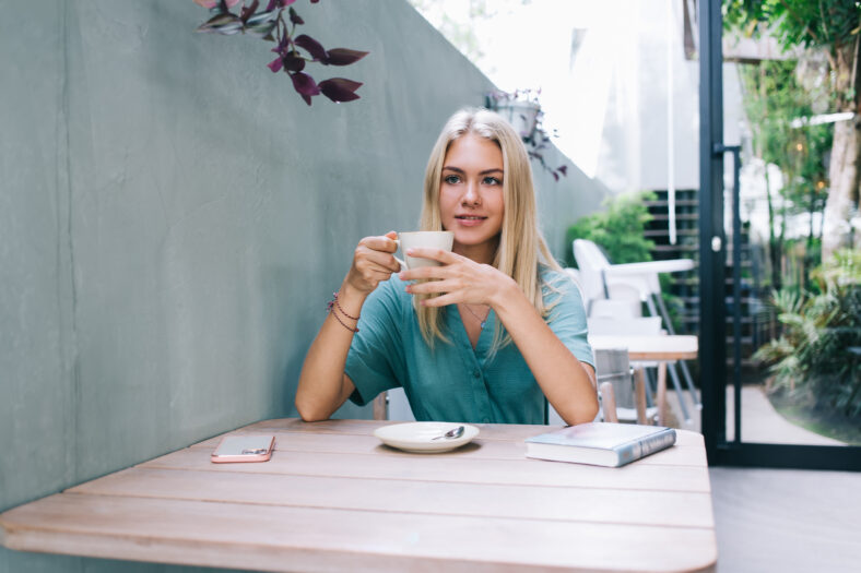 Carefree Caucasian woman with cup of hot caffeine beverage dreaming about leisure spending and positive youth lifestyle, happy blonde female customer resting in outdoors coffee shop enjoying tea time