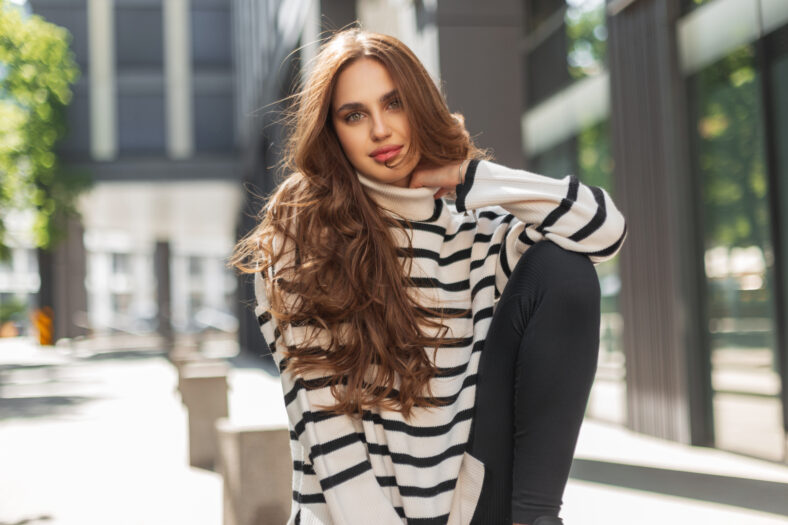Beautiful stylish young woman in a fashionable striped sweater sits and poses on a sunny day in the city