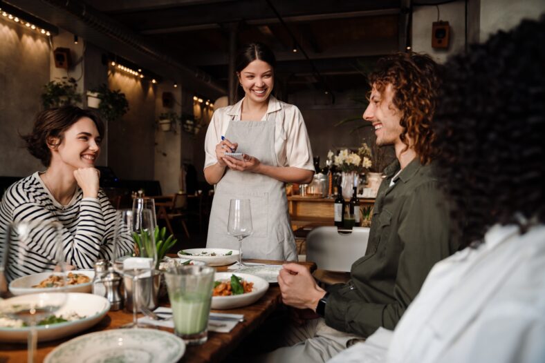 Young smiling waitress wearing apron serving group of cheerful young friends in restaurant