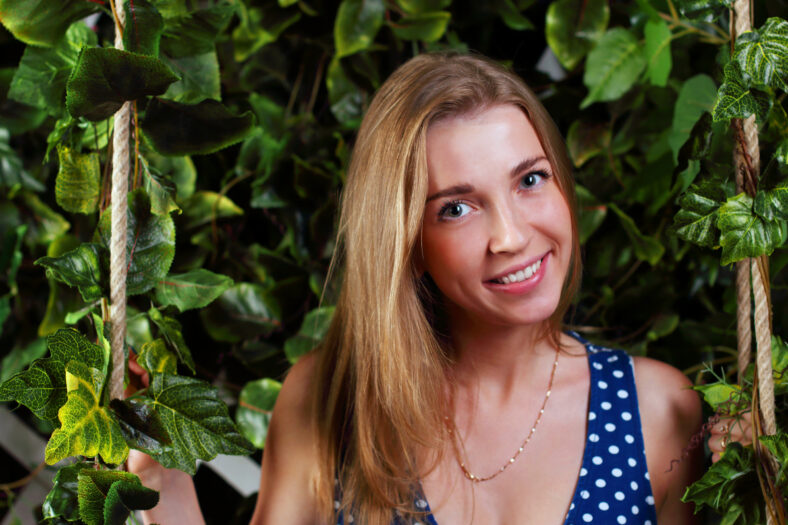 Closeup of beautiful girl with blond hair sitting on swing with green leaves