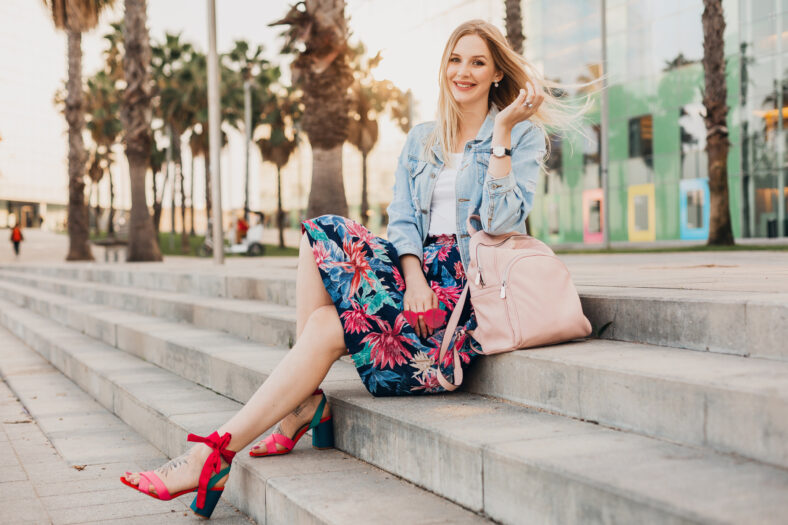 pretty smiling blond woman sitting on stairs in city street in stylish printed skirt and denim oversize jacket with leather backpack, summer style trend