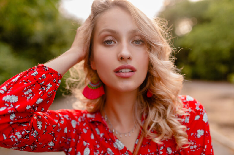 portrait of attractive stylish blond smiling woman in red blouse summer fashion outfit in park boho style wearing earrings smiling white teeth