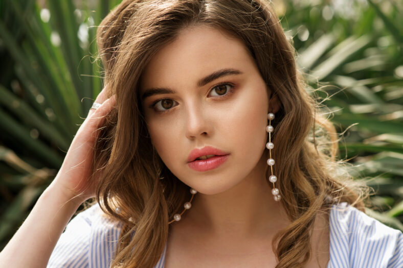 Young beautiful woman with perfect skin and big pearl earrings and curly hair front of greens plants. Close up, selective focus.