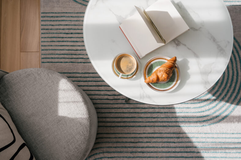 A serene moment captures a marble table featuring a cup of morning coffee, croissant and an open book close to comfortable armchair in living room, flat lay. Slow weekends at home, top view