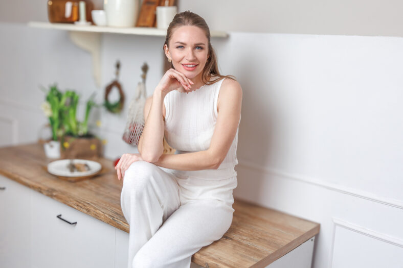 A young woman relaxes on a kitchen counter, showcasing a minimalist interior filled with greenery and natural light, embodying a warm, inviting atmosphere.