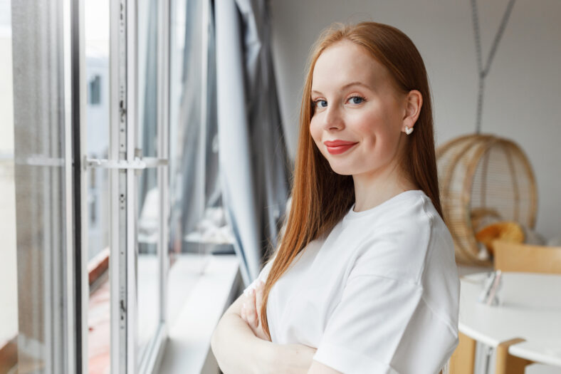 Side view of cute young girl in white t-shirt standing next to big window with folded arms, looking at camera with smile, youthful female student having rest after finishing homework