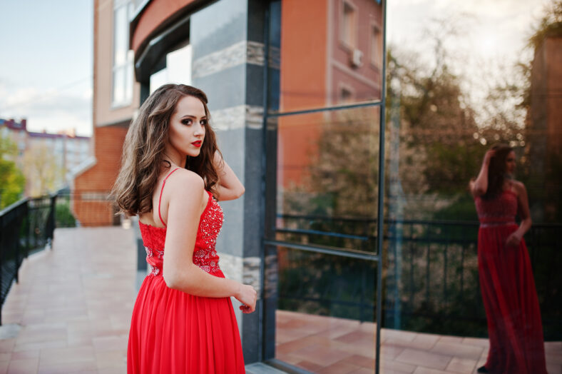 Portrait of fashionable girl at red evening dress posed background mirror window of modern building