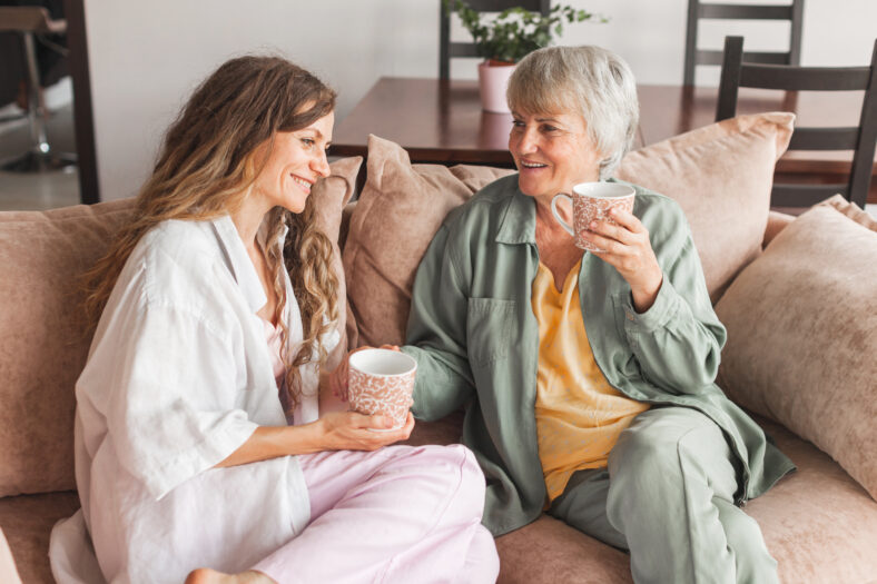 Beautiful mature mother and her adult daughter are drinking coffee, talking and smiling while sitting on couch at home