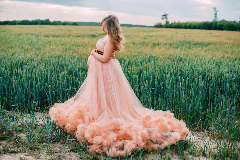 Young happy pregnant woman in peach color cloud dress relaxing and enjoying life in nature. Outdoors on the nature. Pretty pregnant woman holding her tummy. Cloudy dress. Blues sky. Wheat green field.