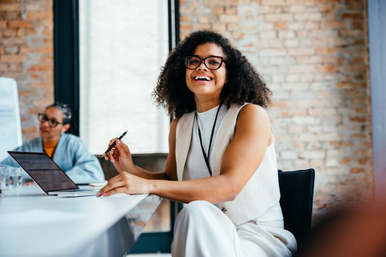 Confident young professional woman smiling in a modern office environment alongside colleague, working on a laptop and engaging in a business meeting.