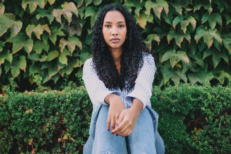 A young woman with beautifully curly hair sits thoughtfully in front of a lush, vibrant green foliage, showcasing a serene and peaceful expression that reflects her calm state of mind