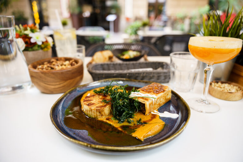 A sophisticated plated meal garnished with leafy greens, served with a rich orange sauce and accompanied by a cocktail in an elegant outdoor restaurant environment