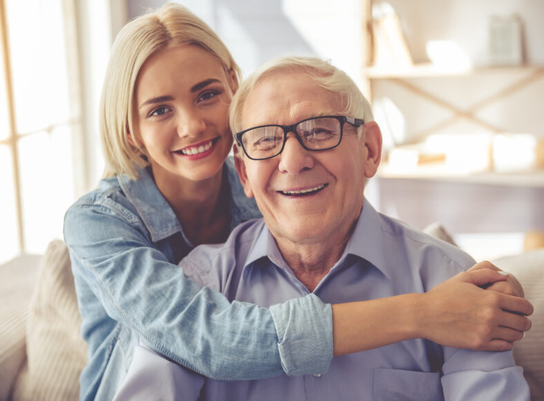 Handsome old man and beautiful young girl are hugging, looking at camera and smiling while sitting on couch at home