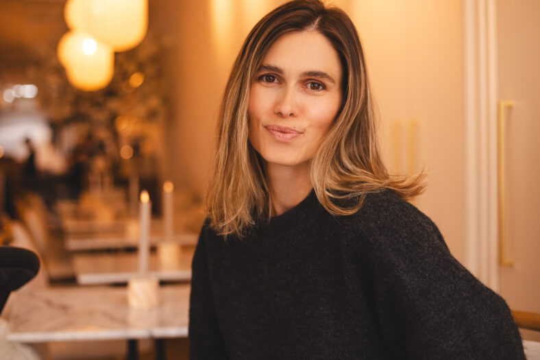 Happy young woman sitting on chair at cafe and looking at camera. Portrait of comfortable woman in wool warm sweater. Portrait of beautiful girl smiling, laughing and relaxing.