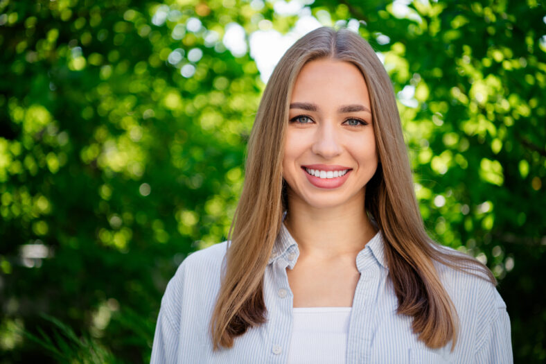 Lovely young woman with a charming smile, enjoying a sunny day outdoors. Surrounded by lush greenery, she embodies youthful energy and happiness in a city park setting.