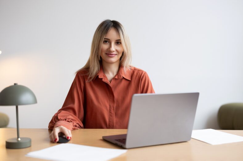 A young woman with long blonde hair sits at a stylish wooden desk, focused on her laptop. Papers are scattered nearby, creating a productive workspace.