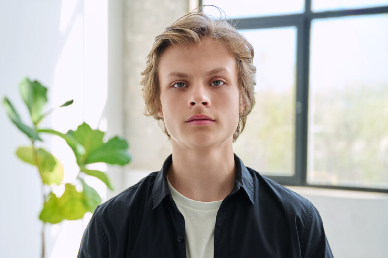 Headshot portrait of serious handsome teenager guy looking at camera, inside home living room. Teenage age, males 16-17 years old, adolescence, education, lifestyle, leisure concept