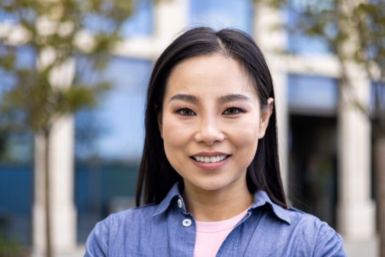 A beautiful asian woman smiles at the camera, her face exuding warmth and confidence, standing outside with a building in the blurred background.