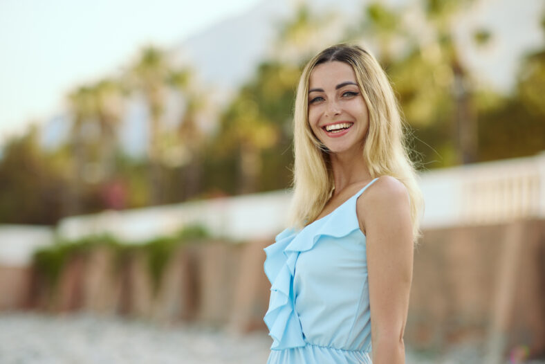 Happy young woman with long blonde hair wearing a ruffled blue dress smiles confidently outdoors near a palm-lined resort, concept of summer joy, travel fashion and lifestyle promo photography.