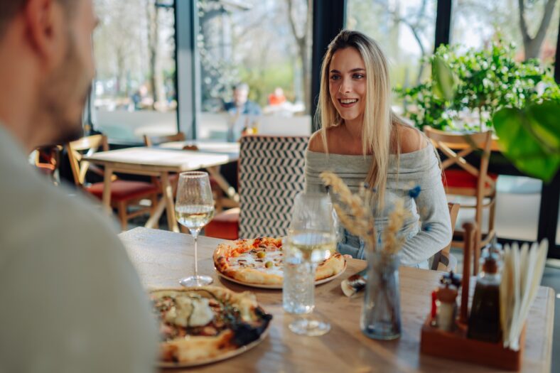 Young woman smiling at her boyfriend while having lunch at a pizzeria, enjoying pizza and white wine
