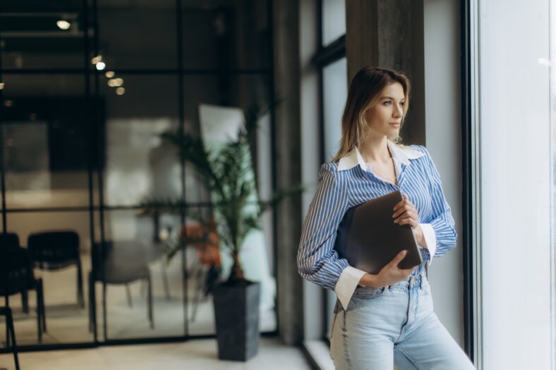 Confident woman standing by an office window holding a laptop, reflecting professionalism. Modern work environment background with contemporary interiors, suggesting focus and productivity.
