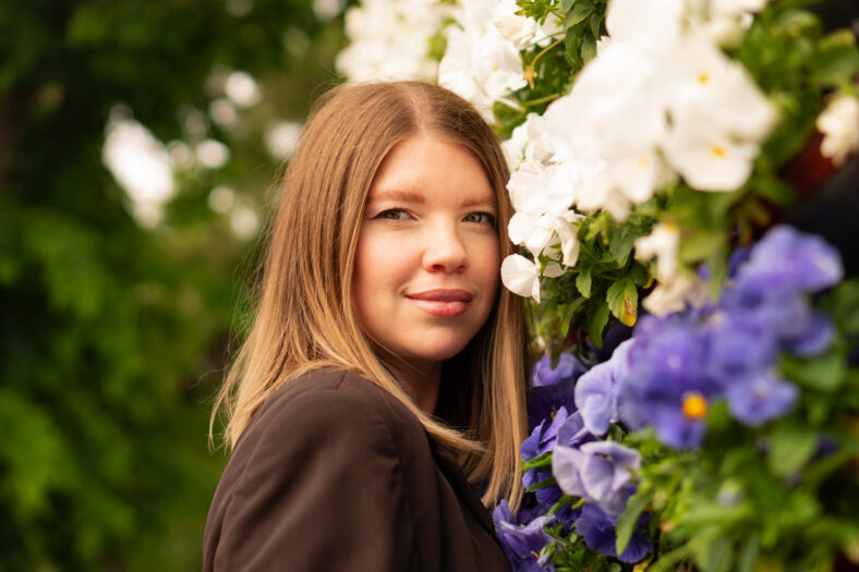 Portrait of smiling young woman beside vibrant floral backdrop outdoors.