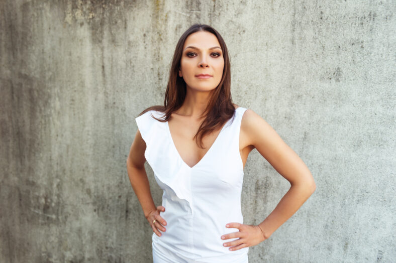 Beautiful slim woman posing outdoors, wearing white dress, standing against grey wall