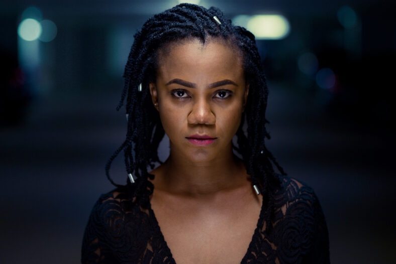 Night portrait of a serious stylish attractive young African woman with braided hair with shining end adornments in a shadowy interior space with defocused lights