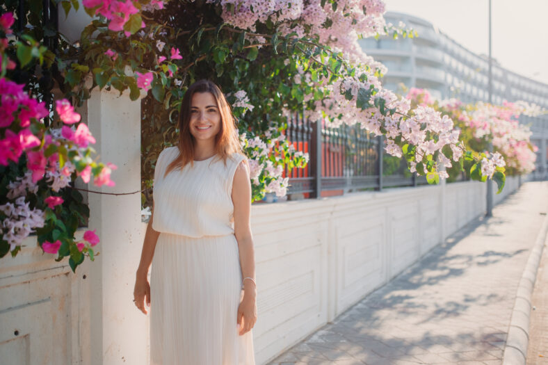 A beautiful brunette girl stands in a white dress on the background of bushes and flowers