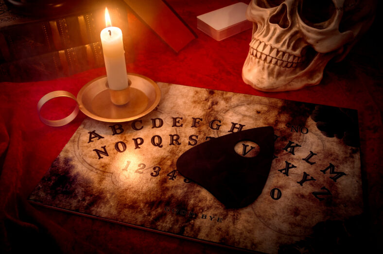 Una guarida de brujas, adivino y concepto de magia negra con un tablero de espíritu parlante iluminado por una vela rodeada de una baraja de cartas, tres libros y un cráneo envuelto con humo misterioso y turbio